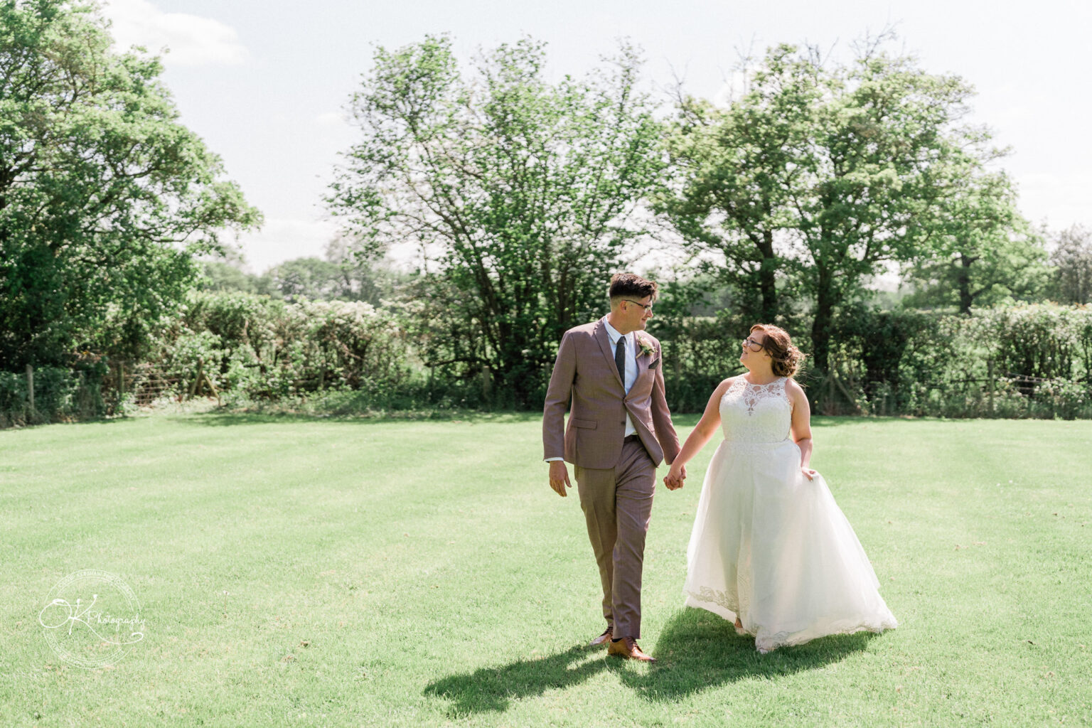 The Old Stables Wedding Photography Newlywed couple holding hands and walking on a green lawn with trees in the background.