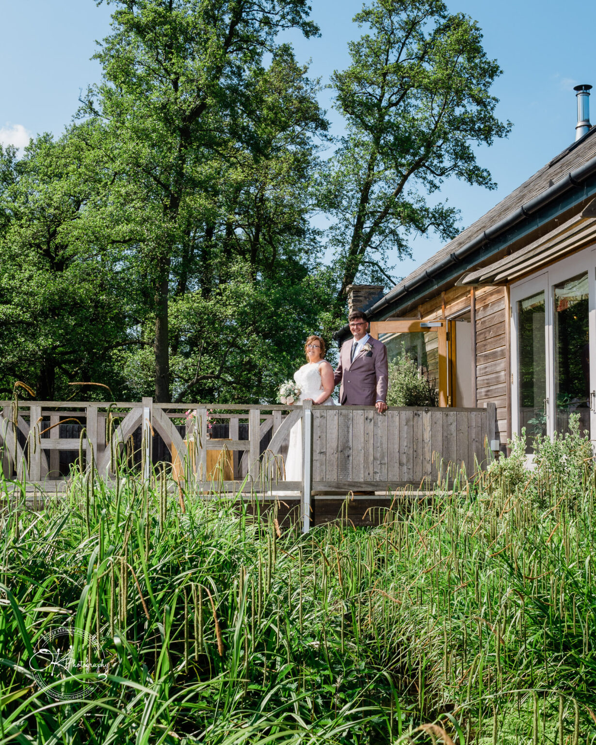 The Old Stables Wedding Photography A bride and groom stand on a wooden deck in front of a rustic building surrounded by greenery.
