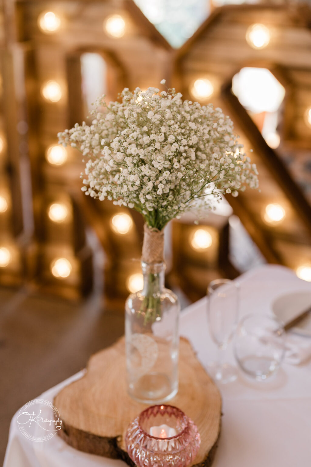 The Old Stables Wedding Photography A wedding table centrepiece featuring a clear bottle with a bouquet of small white flowers, placed on a wooden slice, accompanied by a small candle holder.