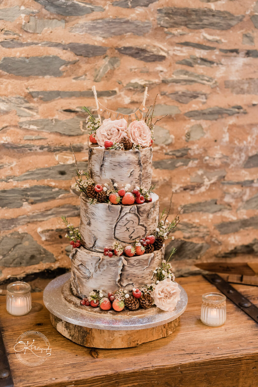 The Old Stables Wedding Photography Three-tiered wedding cake decorated with rustic, birch-inspired design, fresh berries, flowers, and pinecones, placed on a wooden slab.