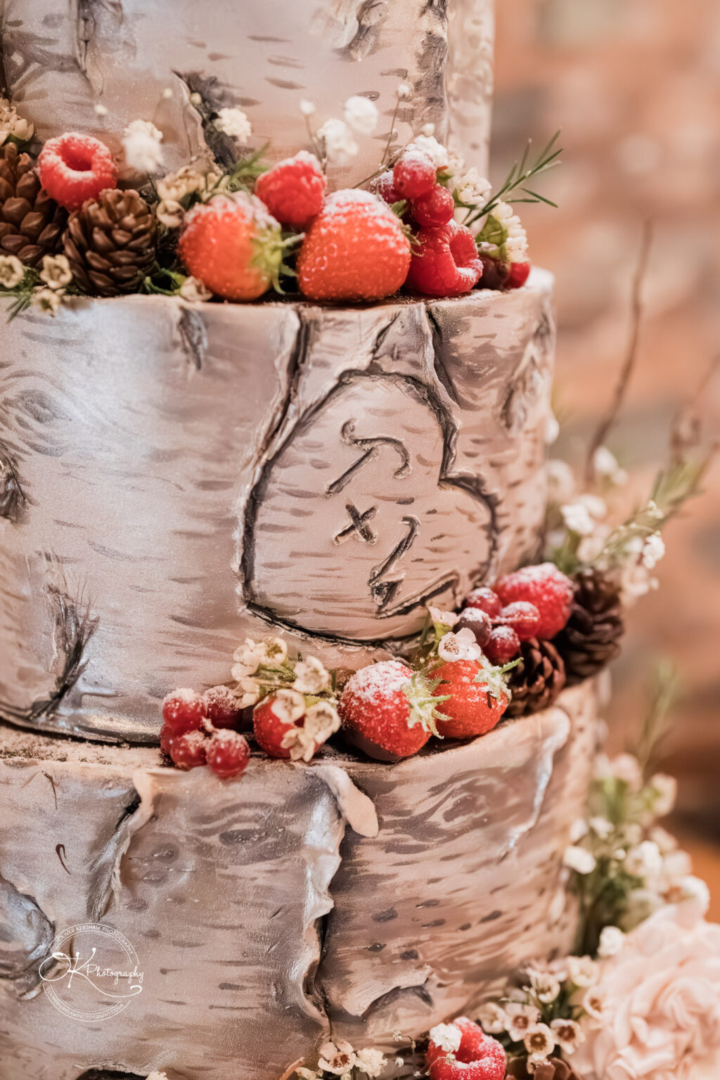 The Old Stables Wedding Photography A close-up of a rustic wedding cake inspired by tree bark, adorned with strawberries, raspberries, white flowers, and pinecones.