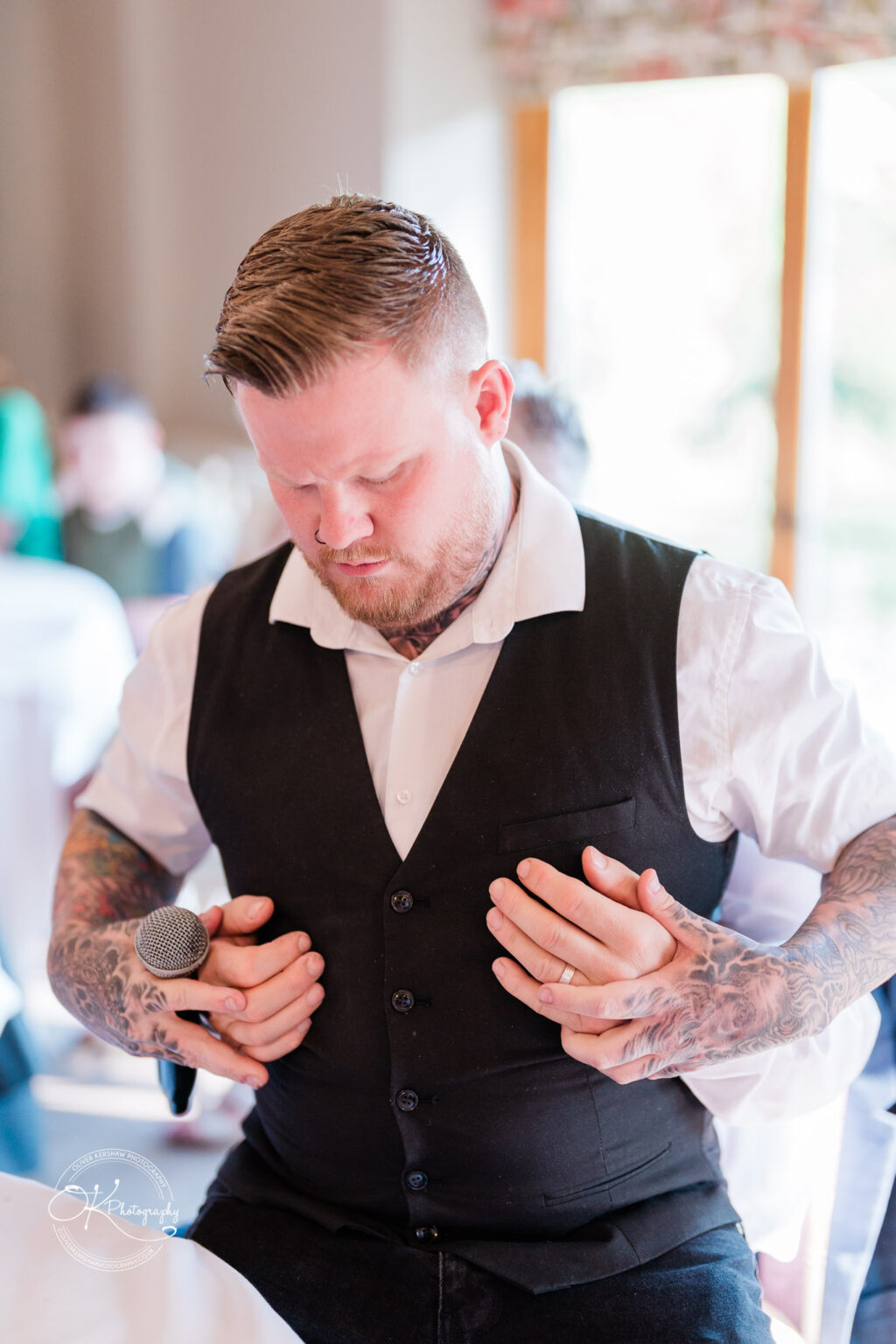 The Old Stables Wedding Photography A man in a black waistcoat and white shirt with tattoos on his arms holds a microphone and places his hands over someone else's hands positioned on his chest during a wedding at The Old Stables.