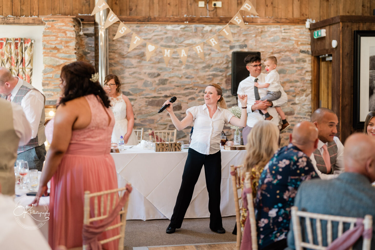 The Old Stables Wedding Photography A lively wedding reception scene with a singer holding a microphone, guests seated at decorated tables, and "Just Married" bunting hanging above.