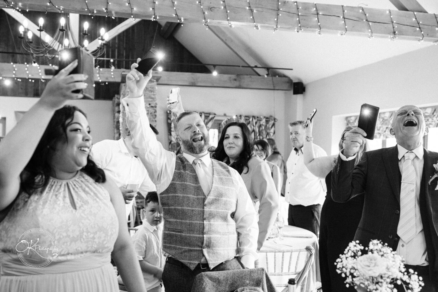 The Old Stables Wedding Photography Guests at a wedding reception cheer while holding up their phones, with visible beams from phone flashlights, in a rustic venue decorated with string lights and exposed wooden beams.