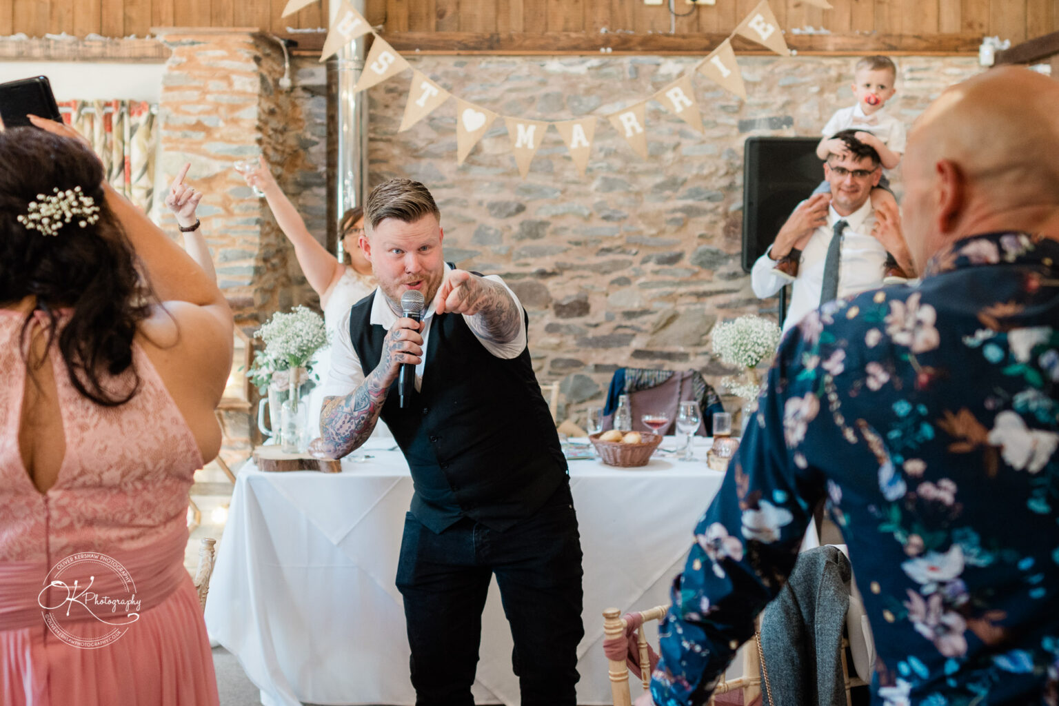 A wedding reception scene with a tattooed man in a vest holding a microphone and pointing, surrounded by guests. A bunting sign saying "JUST MARRIED" hangs in the background, with a bride and groom celebrating.