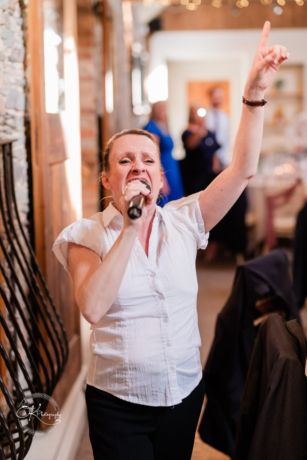 The Old Stables Wedding Photography A woman in a white blouse passionately singing into a microphone with one arm raised, at a wedding venue with stone walls and wooden features.