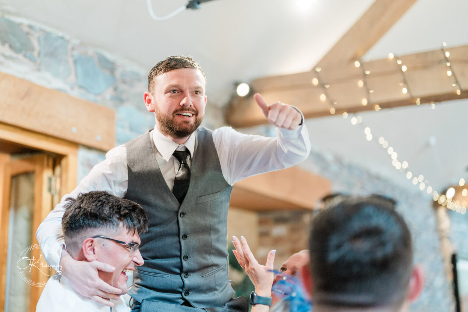 The Old Stables Wedding Photography A man in a grey vest and tie is smiling and giving a thumbs up while being lifted by another man wearing glasses in a rustic venue with stone walls and string lights.