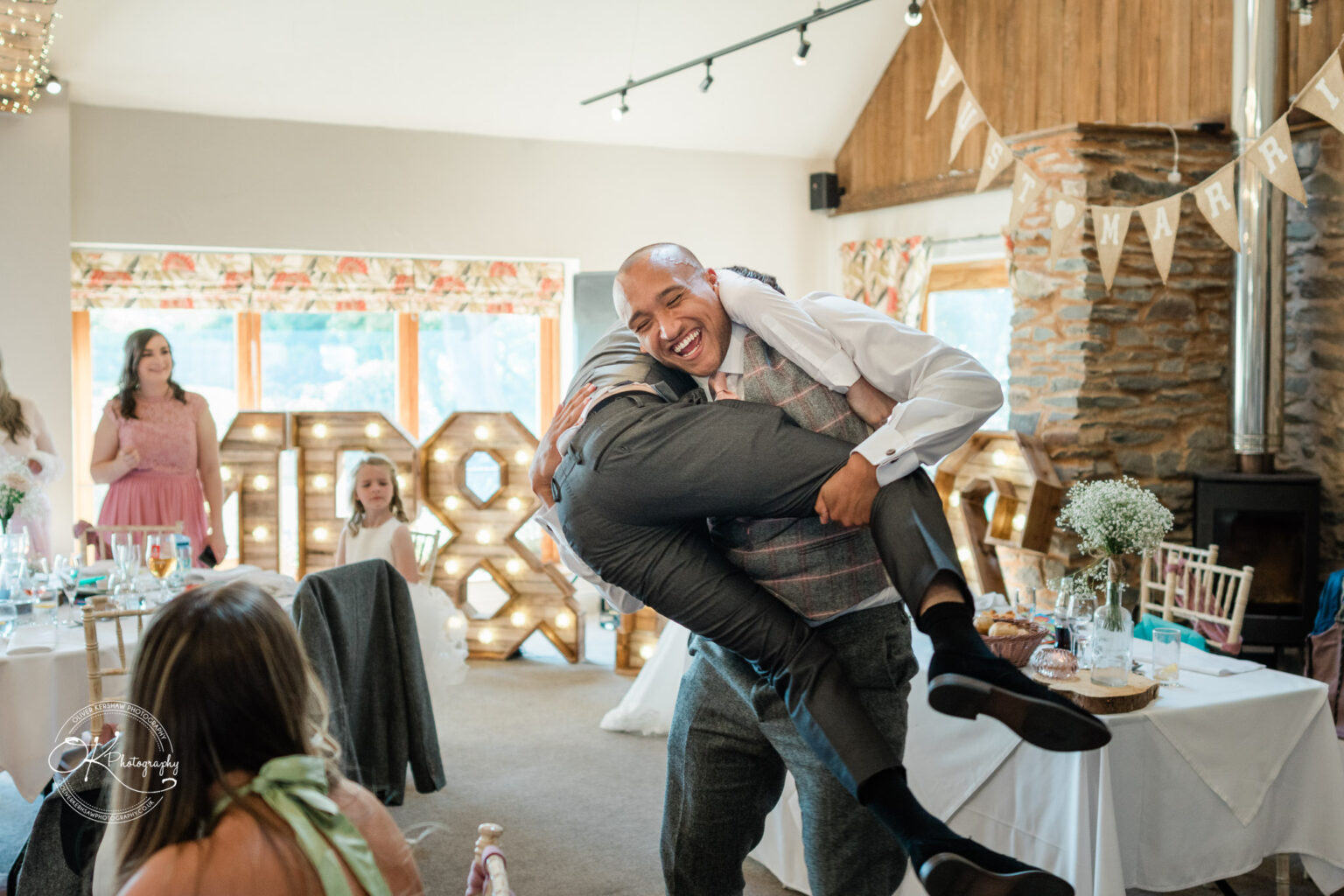 The Old Stables Wedding Photography Guests in a wedding reception hall with rustic decor, including large illuminated initials and bunting. A man in a suit lifts another guest in an embracing gesture with others watching and smiling.