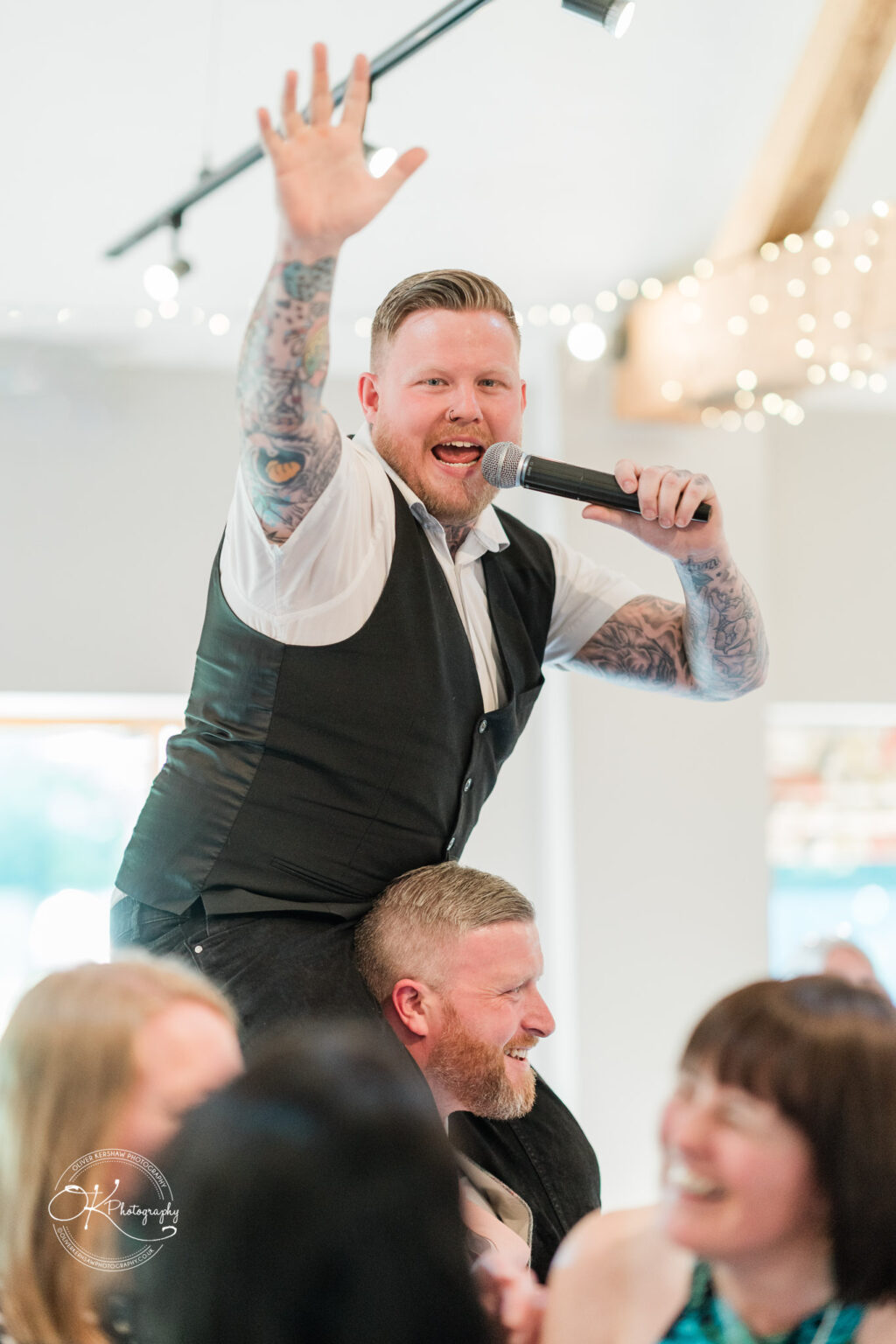 The Old Stables Wedding Photography A man with tattoos holds a microphone and waves while sitting on another man's shoulders at a wedding reception.