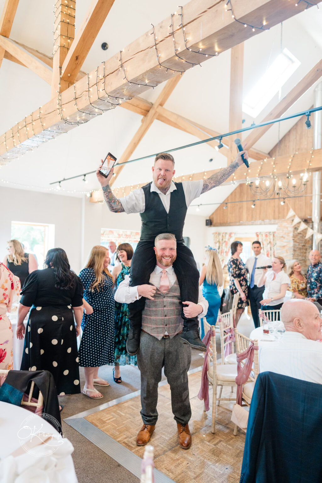 The Old Stables Wedding Photography Man standing on another man’s shoulders, both smiling, amidst guests at a wedding reception in a rustic venue with fairy lights.