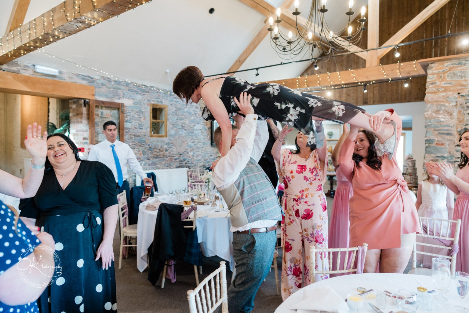 The Old Stables Wedding Photography Guests at a wedding reception joyfully lifting a woman into the air inside a rustic venue with stone walls and wooden beams adorned with fairy lights.
