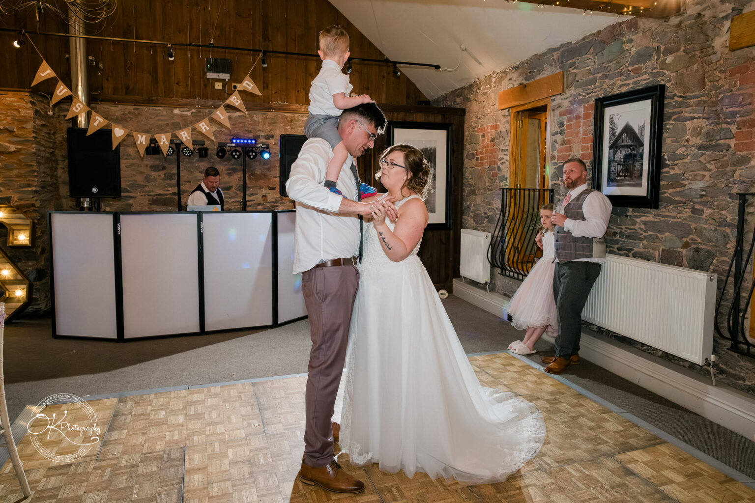 The Old Stables Wedding Photography A bride and groom dance inside a rustic venue with a banner reading "Just Married" hanging in the background. The groom has a child on his shoulders, and there is a DJ booth behind them.