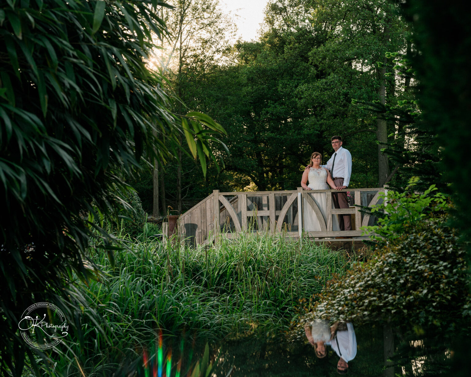 The Old Stables Wedding Photography A wedding couple stands on a wooden bridge surrounded by lush greenery, with their reflection visible in a calm pond below.