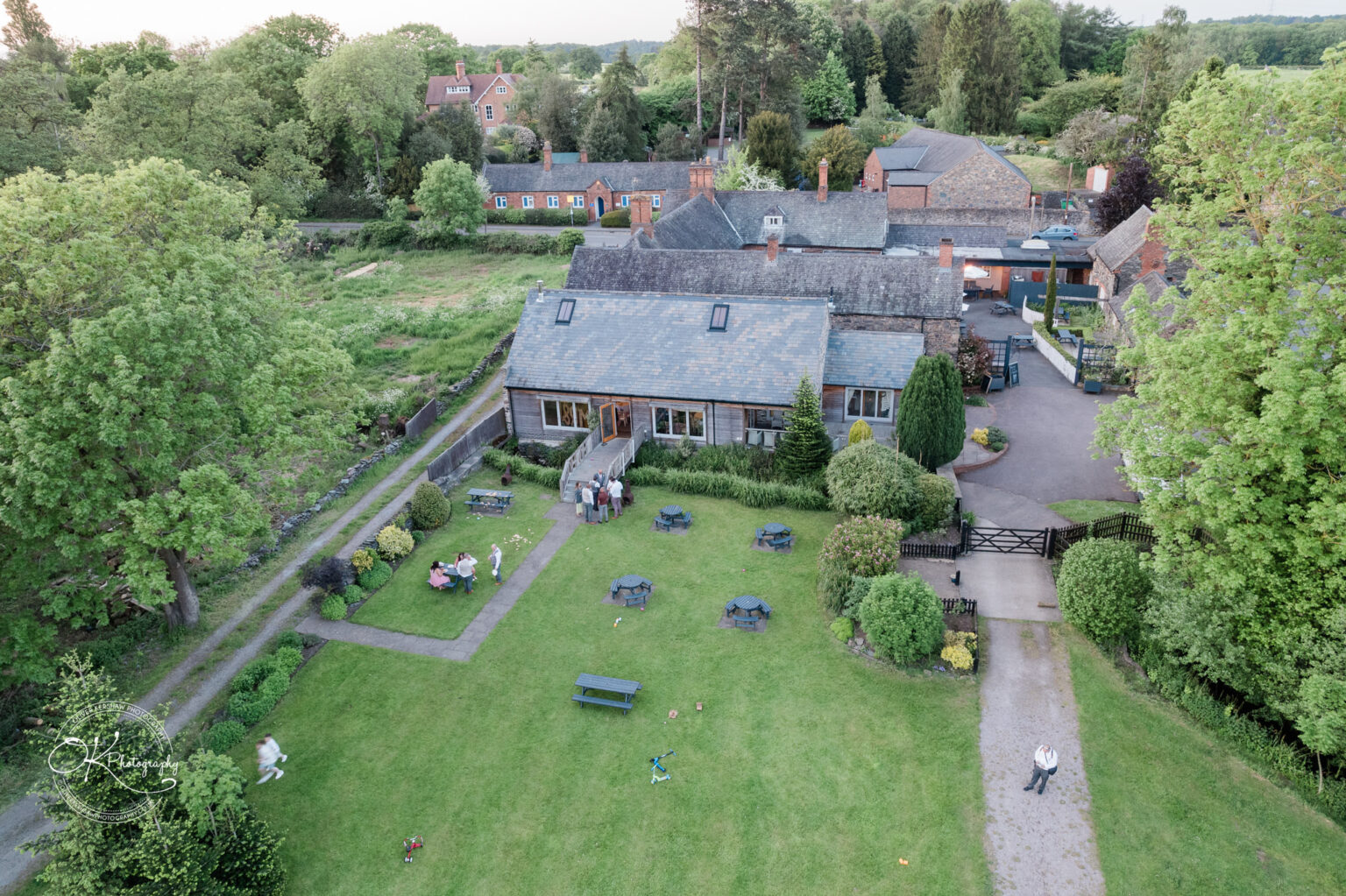 The Old Stables Wedding Photography Aerial view of a country estate with manicured lawns, picnic tables, and a rustic building surrounded by lush greenery, with people gathered outside for a wedding.