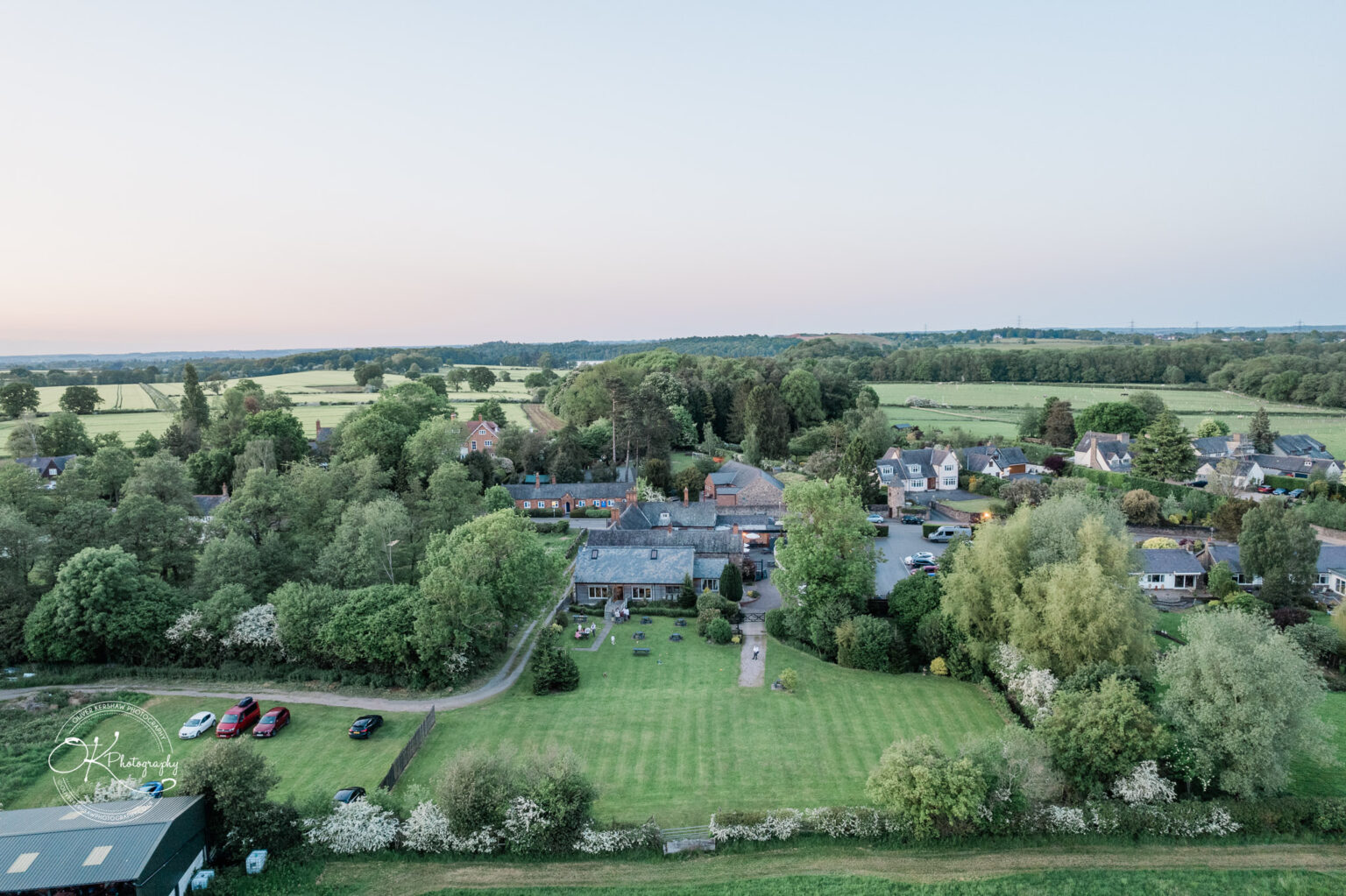 Aerial view of "The Old Stables," showcasing a picturesque venue surrounded by lush greenery, fields, and houses, with a few parked cars on the lawn.