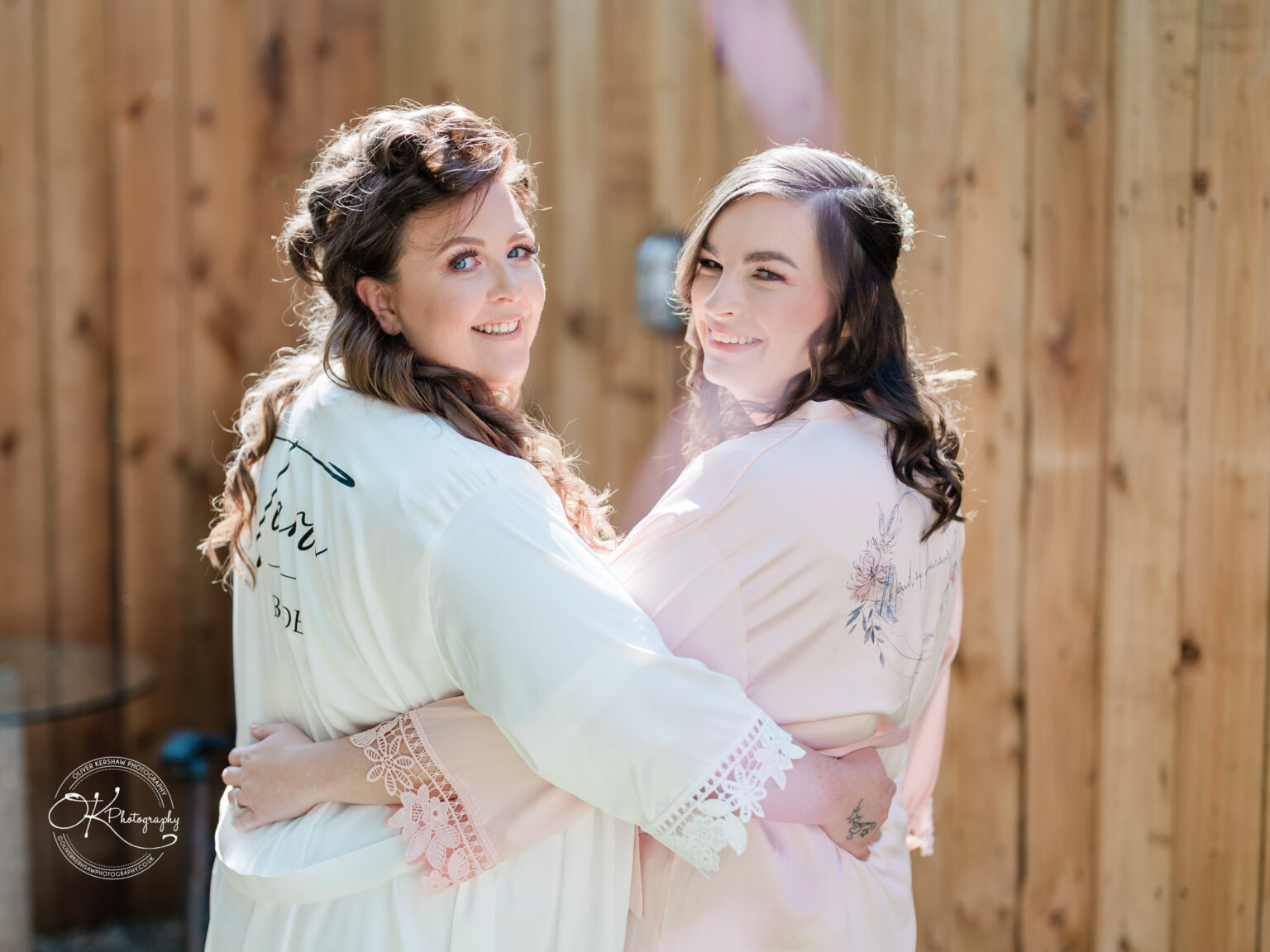 The Old Stables Wedding Photography Two women in robes embrace in front of a wooden fence, smiling at the camera.