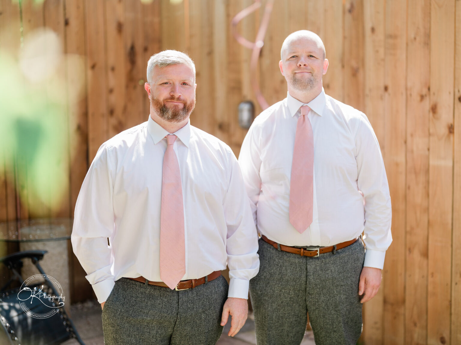 The Old Stables Wedding Photography Two men wearing white shirts, pink ties, and grey trousers, standing in front of a wooden fence.