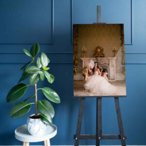 A large framed photograph of a woman in a white gown sits on an easel next to a potted plant against a panelled blue wall.