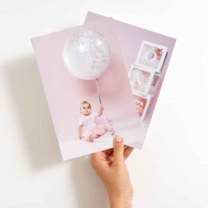 Hand holding two photographs, one featuring a baby sitting with a balloon and the other showing "ONE" white block letters against a pink background.