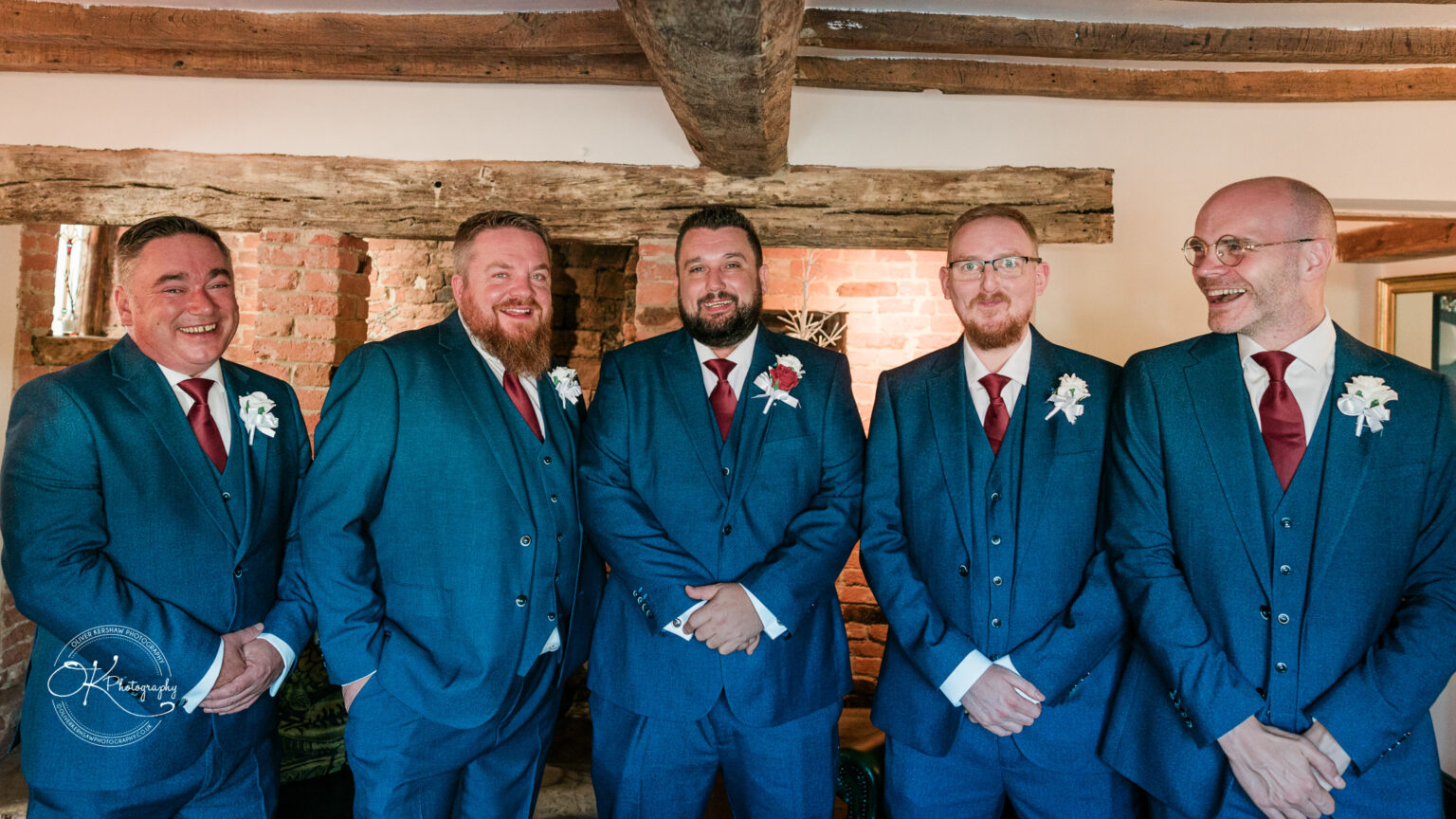 The cottage hotel - Wedding Photography Five men in blue suits and red ties stand together smiling, beneath a wooden beam in a rustic room with exposed brick walls.