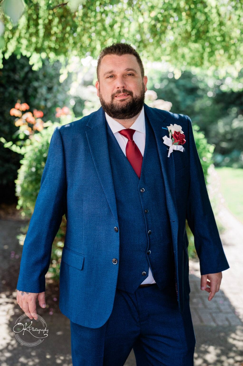 The cottage hotel - Wedding Photography A man in a blue suit with a red tie and floral boutonniere standing outdoors under greenery.
