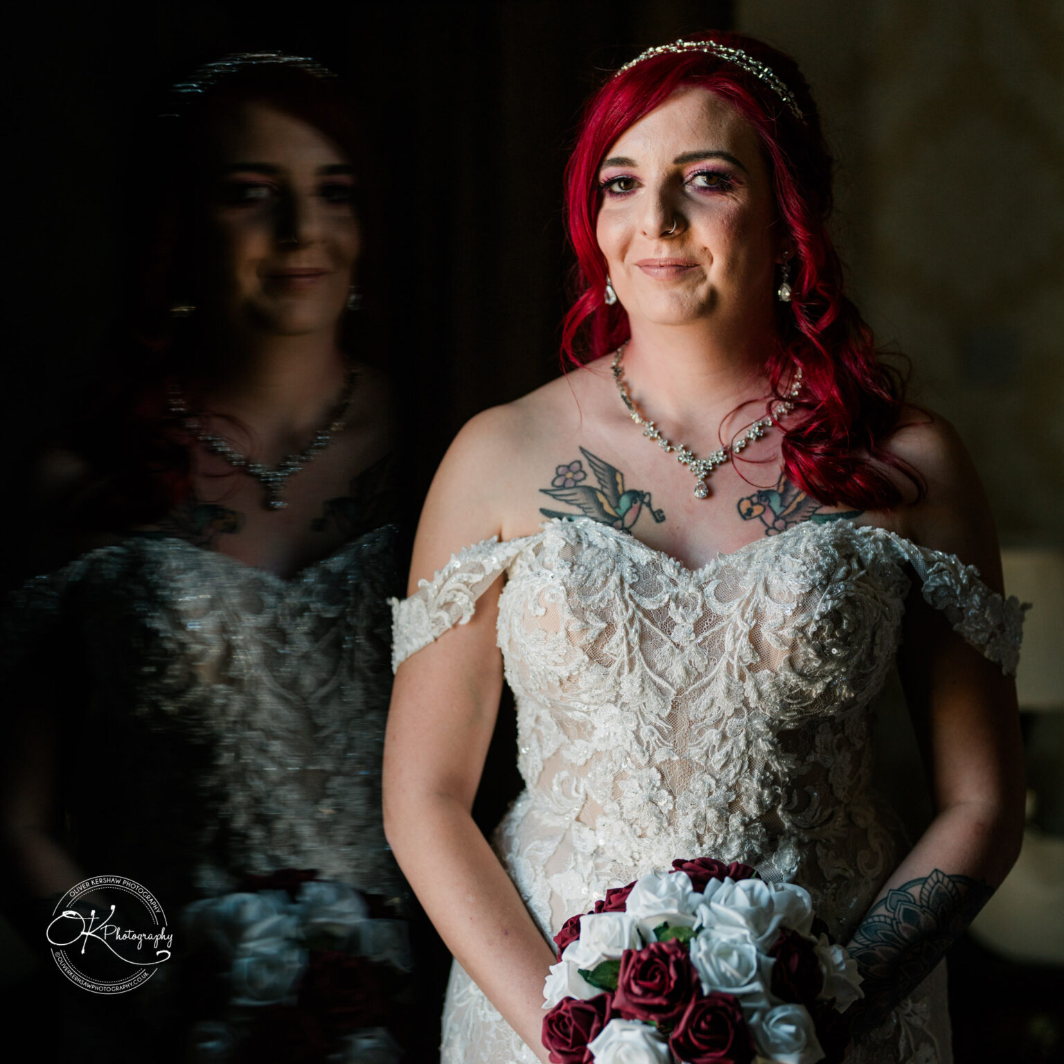 The cottage hotel - Wedding Photography Bride with red hair in a lace wedding dress holding a bouquet of red and white roses, standing next to a mirror reflecting her image.