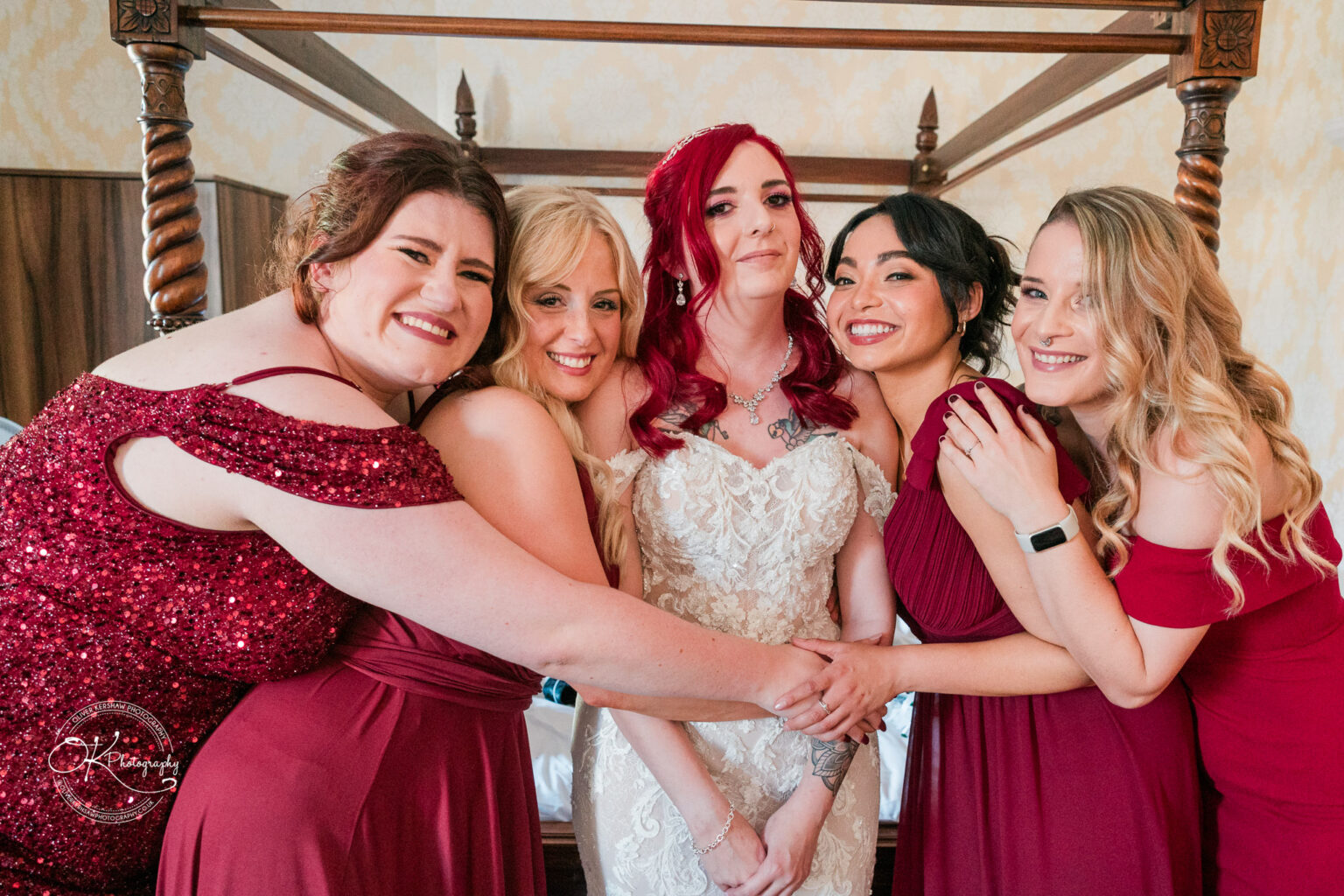 The cottage hotel - Wedding Photography A bride in a white gown is surrounded by four women dressed in red dresses, all smiling and embracing each other in a hotel room.