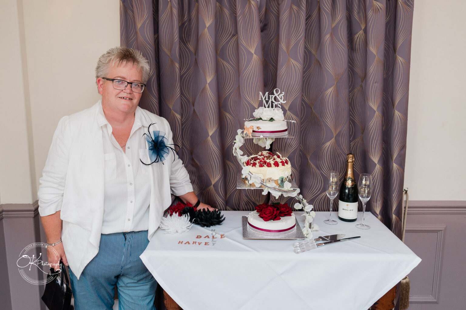 The cottage hotel - Wedding Photography A person standing next to a three-tiered wedding cake and a bottle of champagne on a table, with purple curtains in the background.