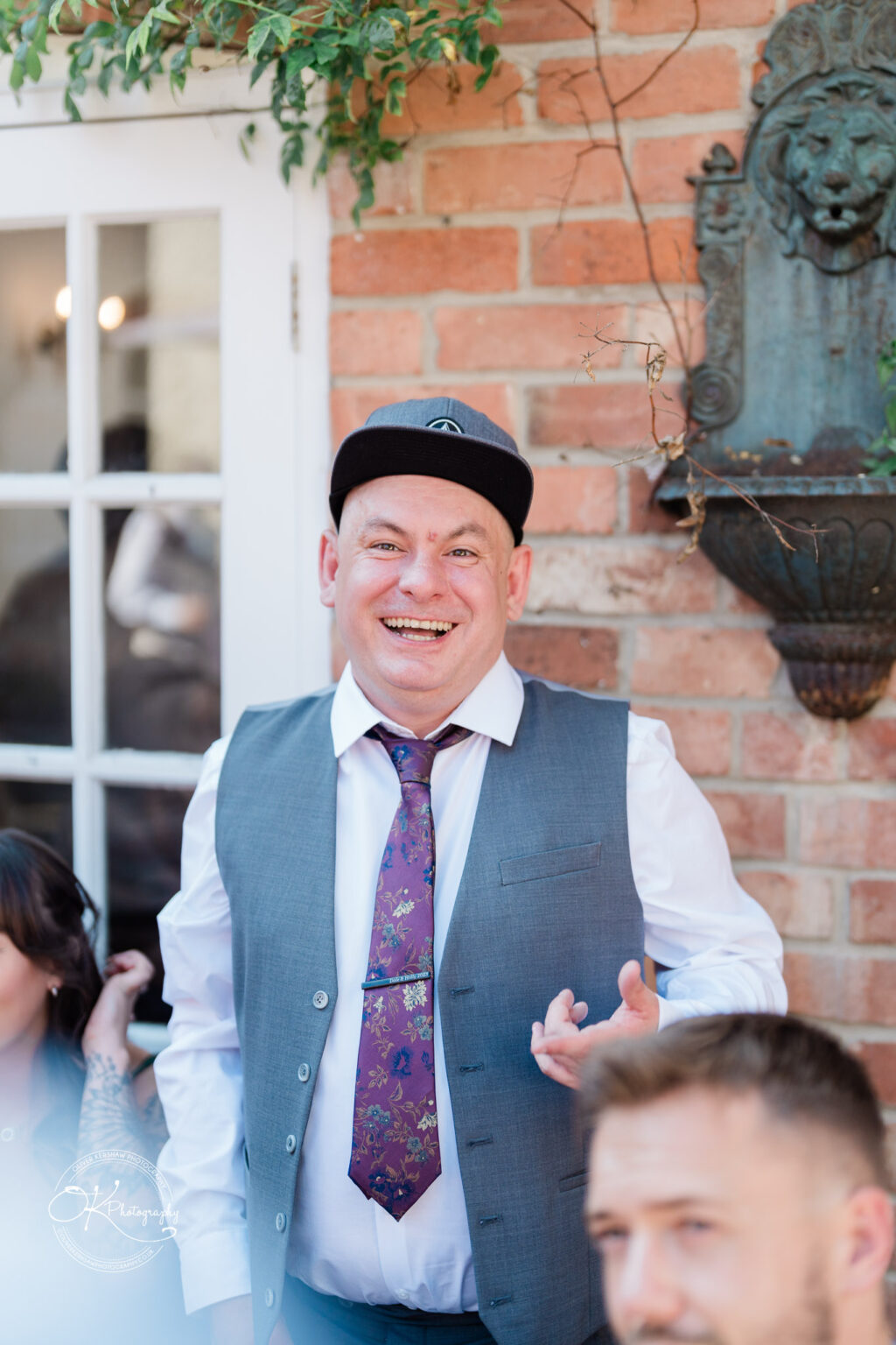 The cottage hotel - Wedding Photography A man in formal attire, wearing a grey waistcoat, white shirt, floral tie, and a black cap, smiles while standing in front of a brick wall and a window with white frames. There is a decorative element on the wall and some greenery above the window.