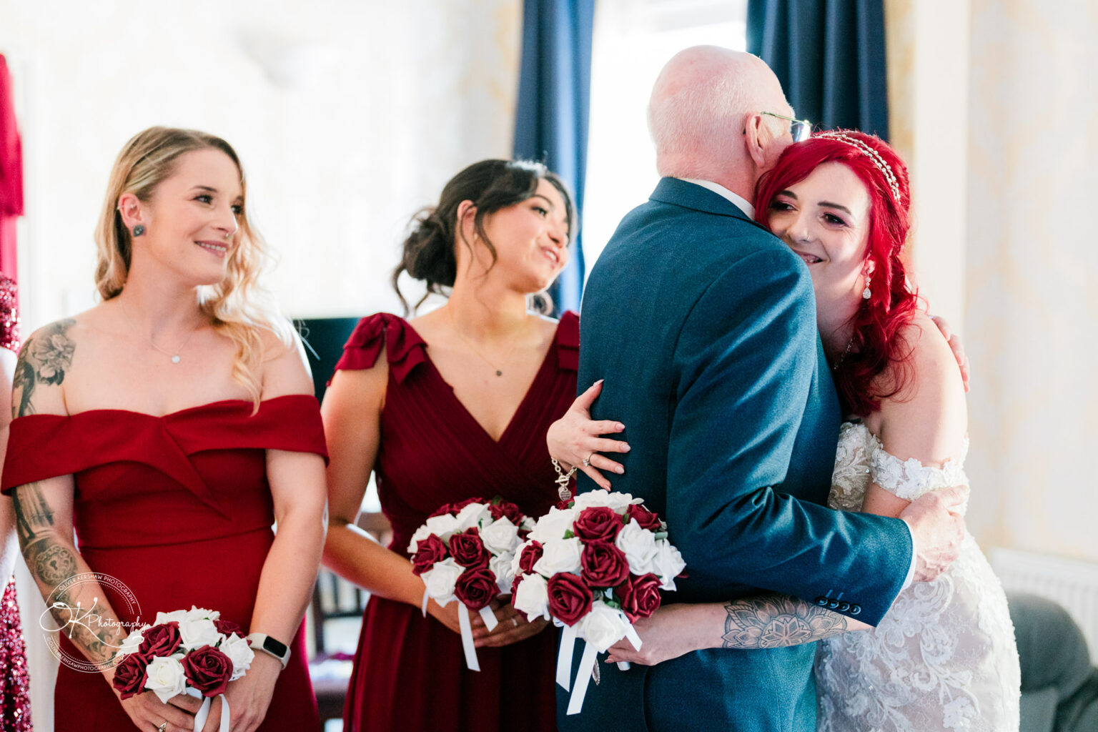 The cottage hotel - Wedding Photography A wedding scene with a bride in a white dress hugging an older man dressed in a suit. Two bridesmaids in red dresses holding bouquets stand to the side, smiling.