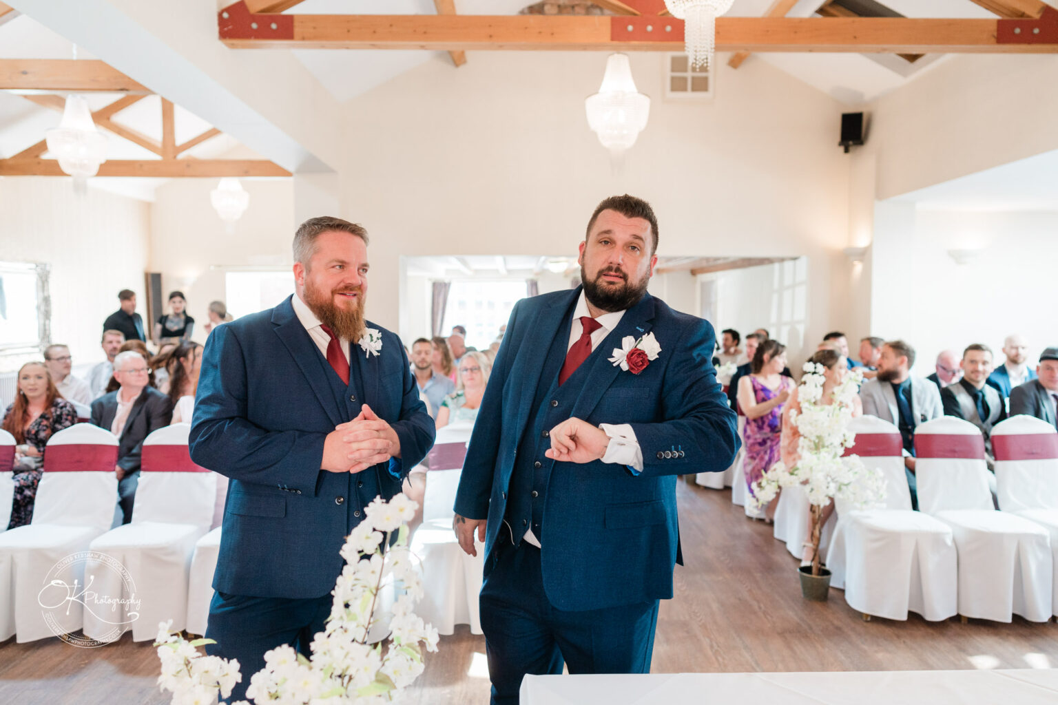 The cottage hotel - Wedding Photography Two groomsmen in blue suits and red ties stand in an indoor wedding venue, with guests seated in the background.
