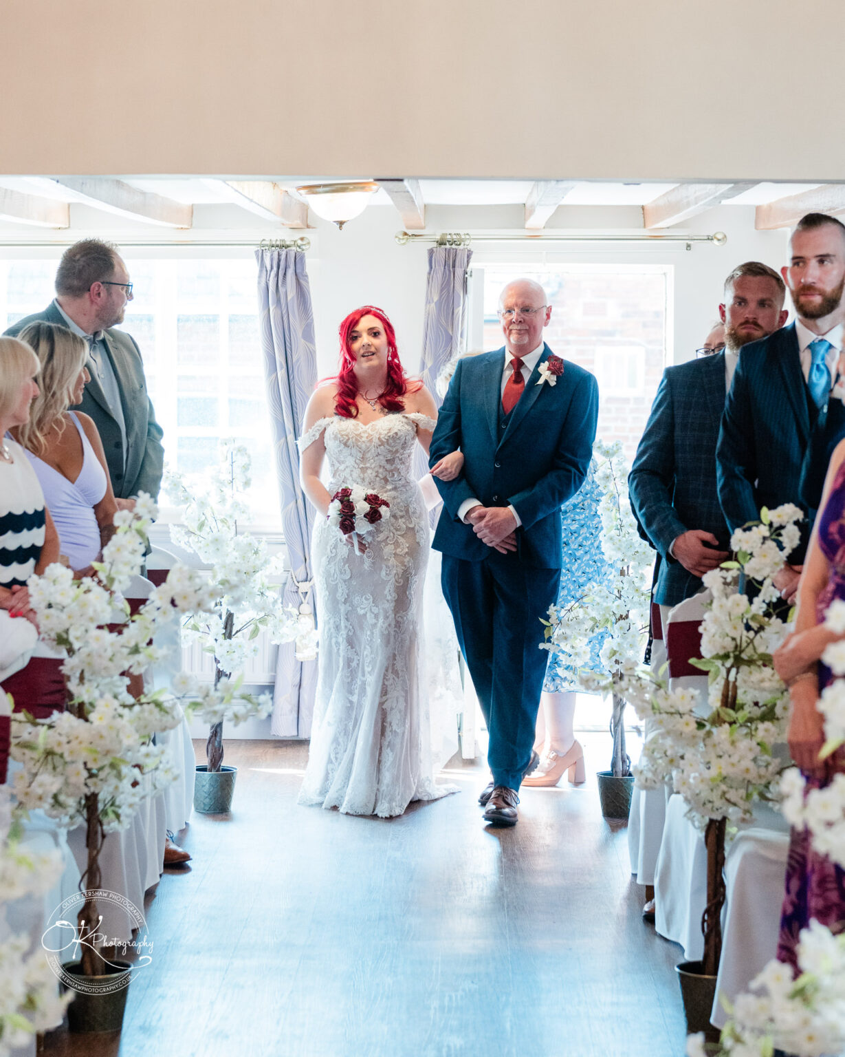 The cottage hotel - Wedding Photography Bride in off-shoulder lace wedding dress and holding a bouquet of red roses, walking down the aisle arm in arm with an older man in a blue suit, with seated guests on both sides and white flower decorations lining the aisle.