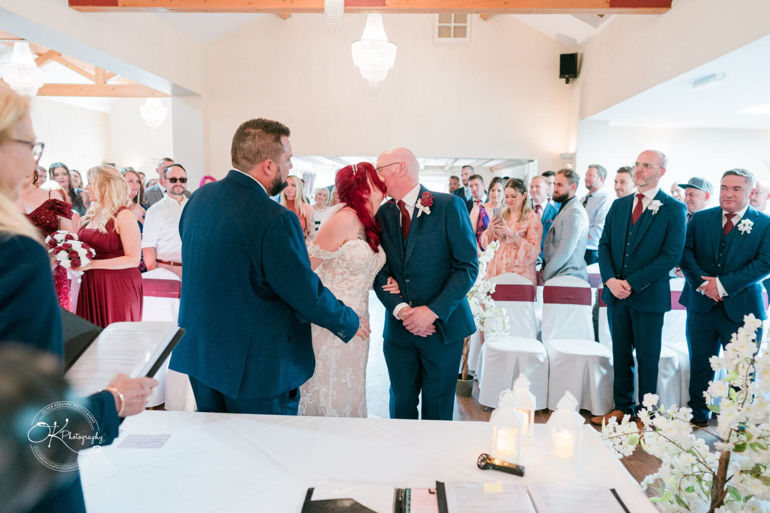 The cottage hotel - Wedding Photography A wedding ceremony with a bride in a white dress kissing an elderly man, a man in a suit stands next to them, and guests are seated and standing in the background.