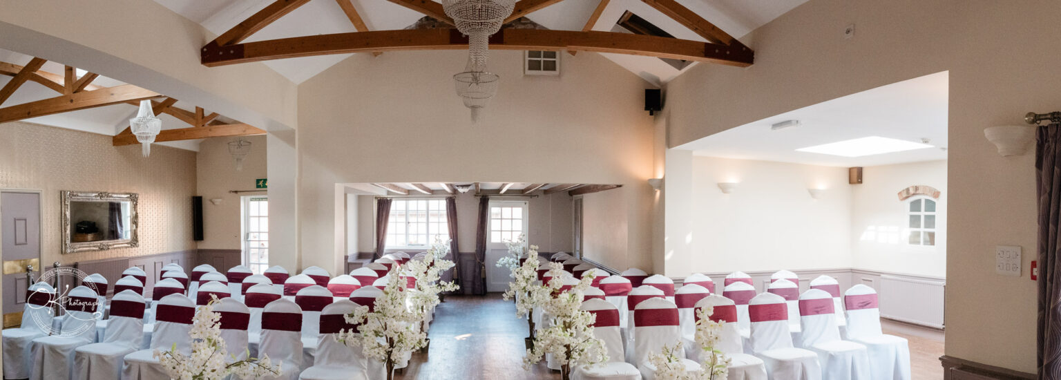 The cottage hotel - Wedding Photography Conference room set up with rows of chairs draped in white covers and adorned with maroon sashes, a chandelier hanging from the ceiling, and decorative flowers lining the central aisle.