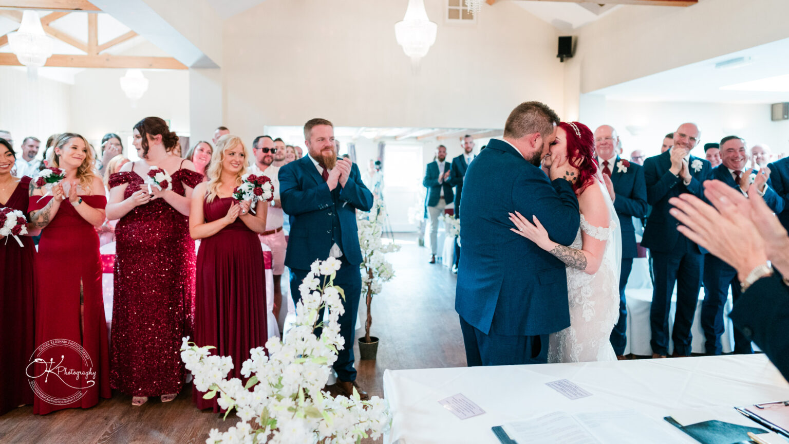 The cottage hotel - Wedding Photography A newlywed couple kissing at their wedding ceremony while guests, including bridesmaids in red dresses and groomsmen in suits, applaud.