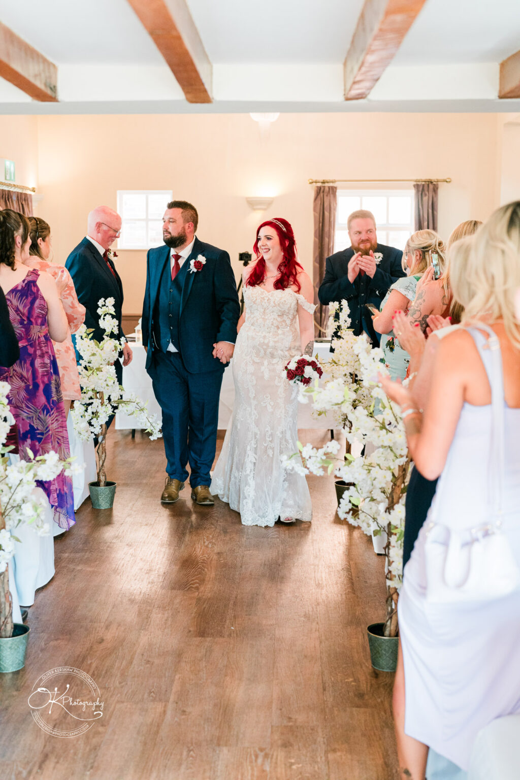 The cottage hotel - Wedding Photography Newly married couple holding hands, walking down the aisle while guests clap and smile inside a decorated room at The Cottage Hotel.