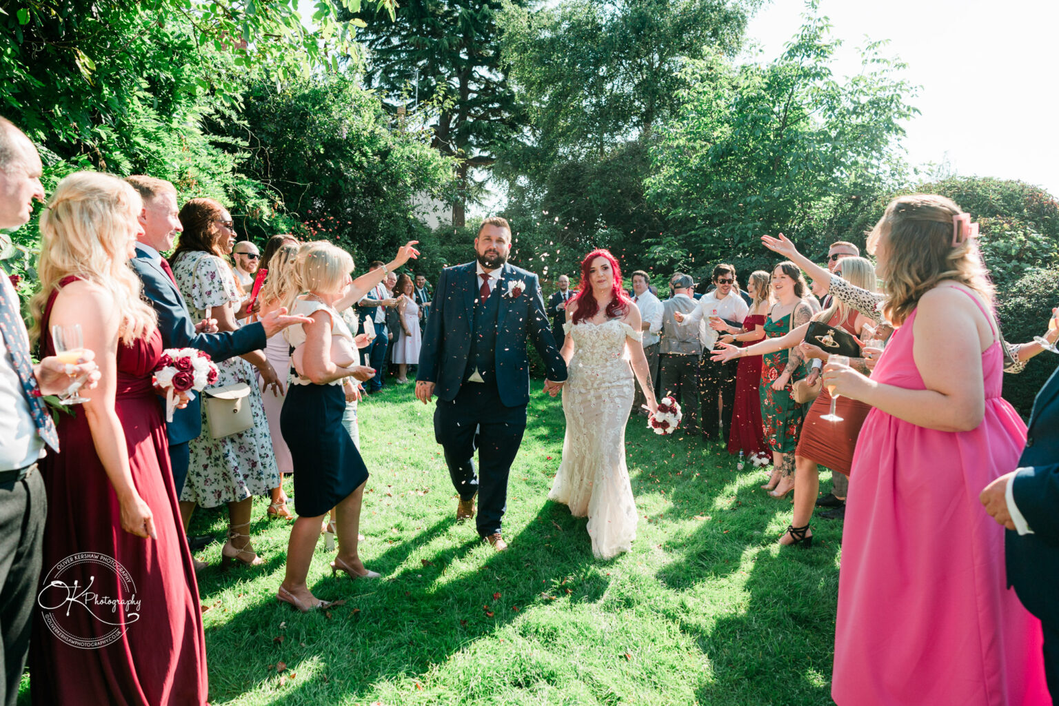 The cottage hotel - Wedding Photography A bride and groom walk hand in hand down a grassy aisle outside, surrounded by guests throwing confetti.
