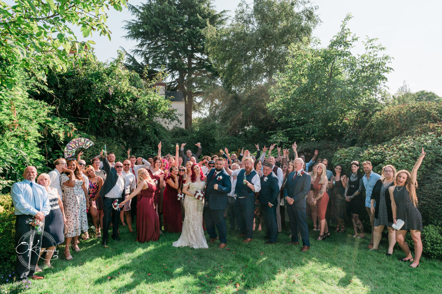 The cottage hotel - Wedding Photography A large group of people, dressed in formal attire, posing outdoors in a lush garden for a celebratory group photo.