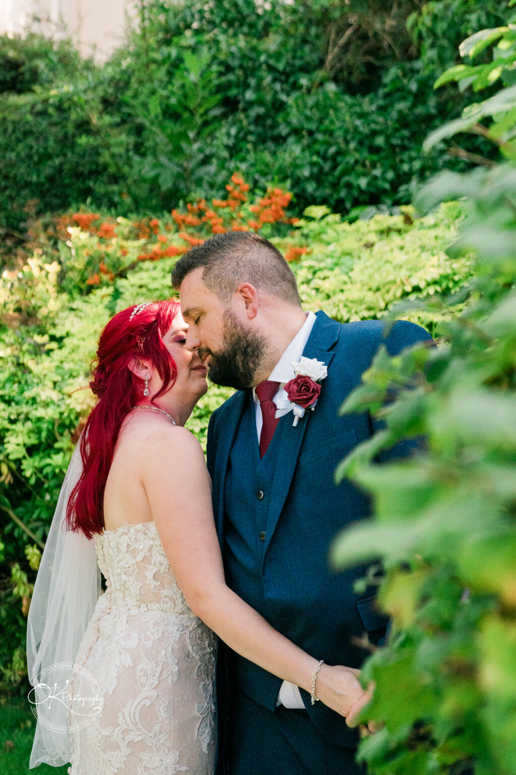 The cottage hotel - Wedding Photography Bride and groom sharing a tender moment in a lush garden with vibrant green foliage and orange flowers in the background.