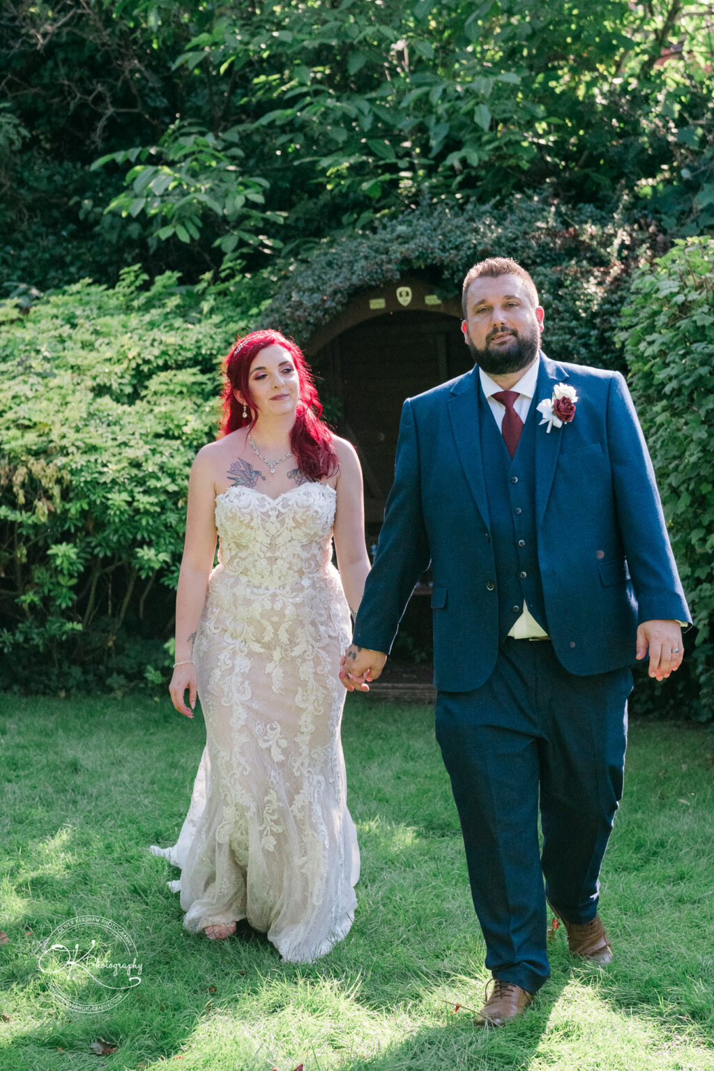 The cottage hotel - Wedding Photography Bride with red hair in a lace wedding dress and groom in a blue suit holding hands outdoors, with lush greenery in the background.