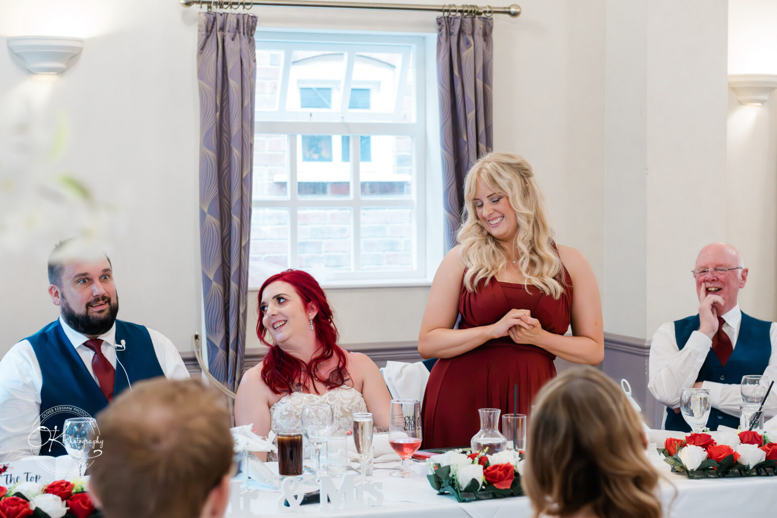 The cottage hotel - Wedding Photography A wedding reception scene with guests seated at a table decorated with red and white flowers. A woman in a red dress is standing and giving a speech, while others around her are smiling and laughing.