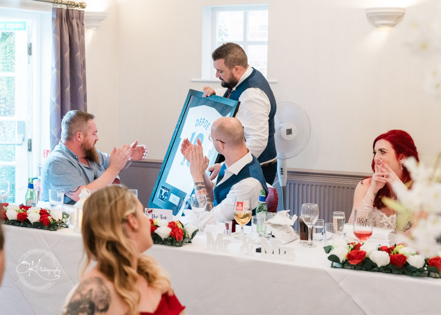 The cottage hotel - Wedding Photography People sitting at a decorated table in a hotel, applauding a man holding a framed football shirt.