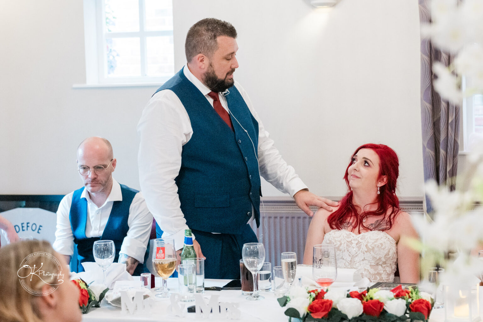 The cottage hotel - Wedding Photography A man in a blue waistcoat is giving a speech while a woman with red hair in a white dress looks at him, with another man seated at a table in the background.