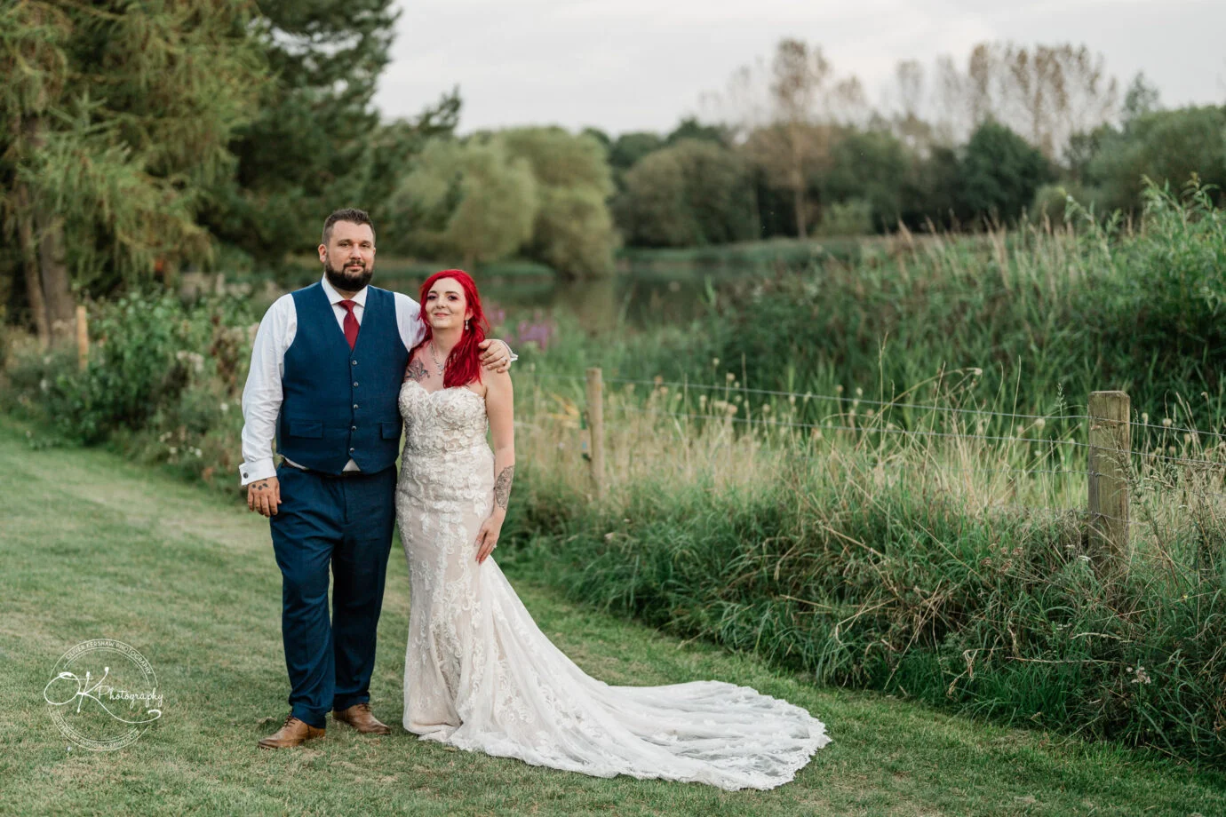 A couple in formal attire, with the man in a vest and the woman in a wedding dress, standing in a grassy outdoor setting with trees and a lake in the background.