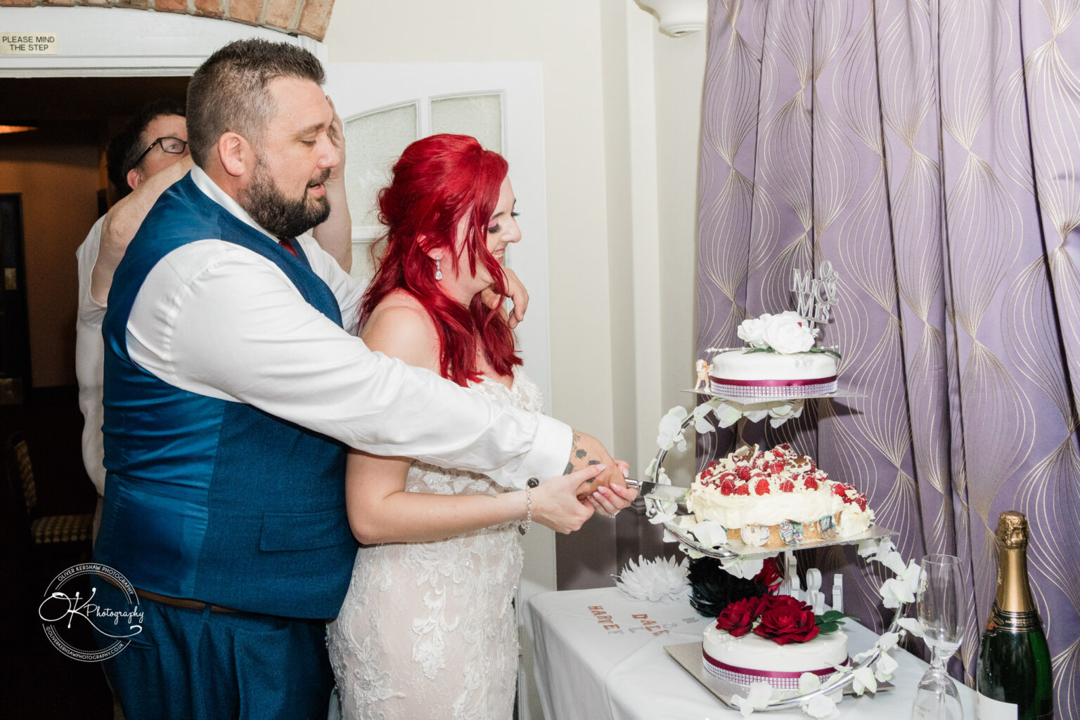 The cottage hotel - Wedding Photography A bride and groom cutting their wedding cake, with the groom standing behind the bride, holding her hand. The tiered cake is decorated with flowers and a "Mr & Mrs" topper.