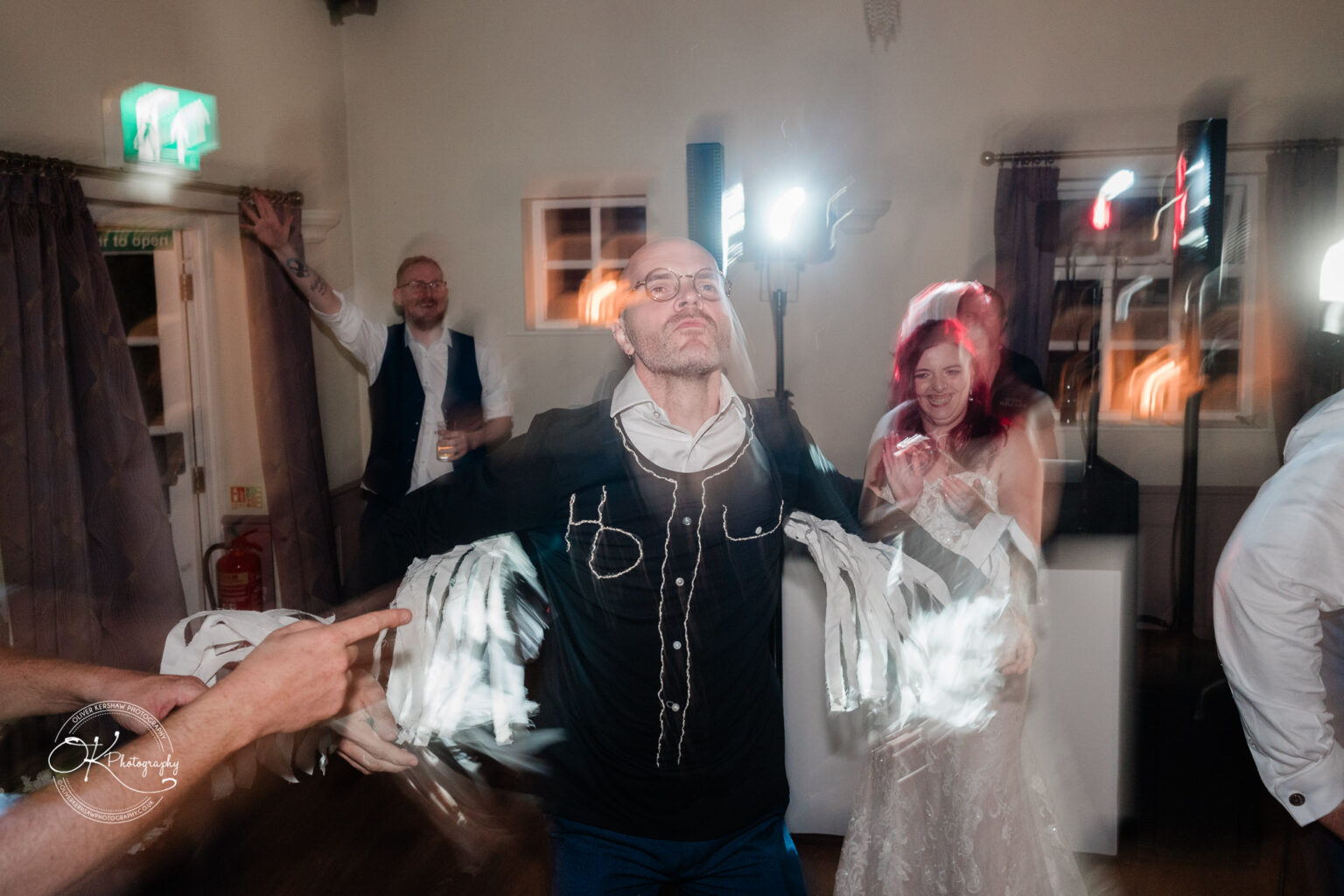 The cottage hotel - Wedding Photography People dancing and celebrating in a room with dim lighting, a man in the foreground with arms raised wearing glasses and a costume, others in the background laughing and clapping.