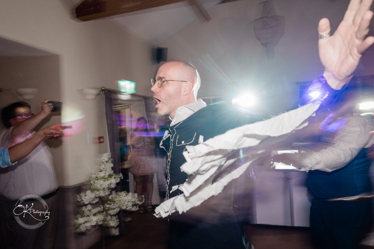 The cottage hotel - Wedding Photography A man with glasses and an open mouth is dancing energetically indoors, with motion blur and light streaks capturing the movement. Others are seen in the background, one holding a camera.