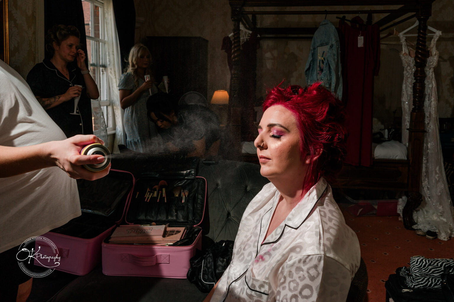 The cottage hotel - Wedding Photography A woman with red hair sits with her eyes closed while a makeup artist sprays her face, surrounded by beauty products and onlookers in a cosy hotel room.