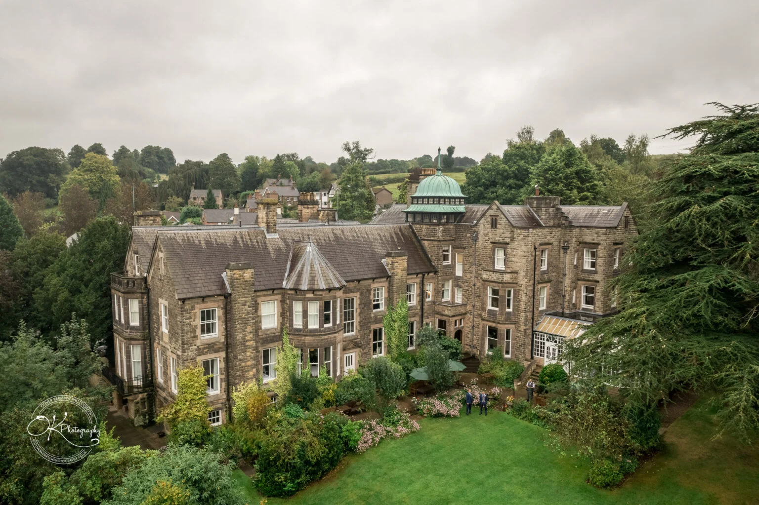 Aerial view of the Makeney Hall Hotel, a large stone building surrounded by lush green gardens and trees, with a dome structure on the roof and people gathered on the lawn below.