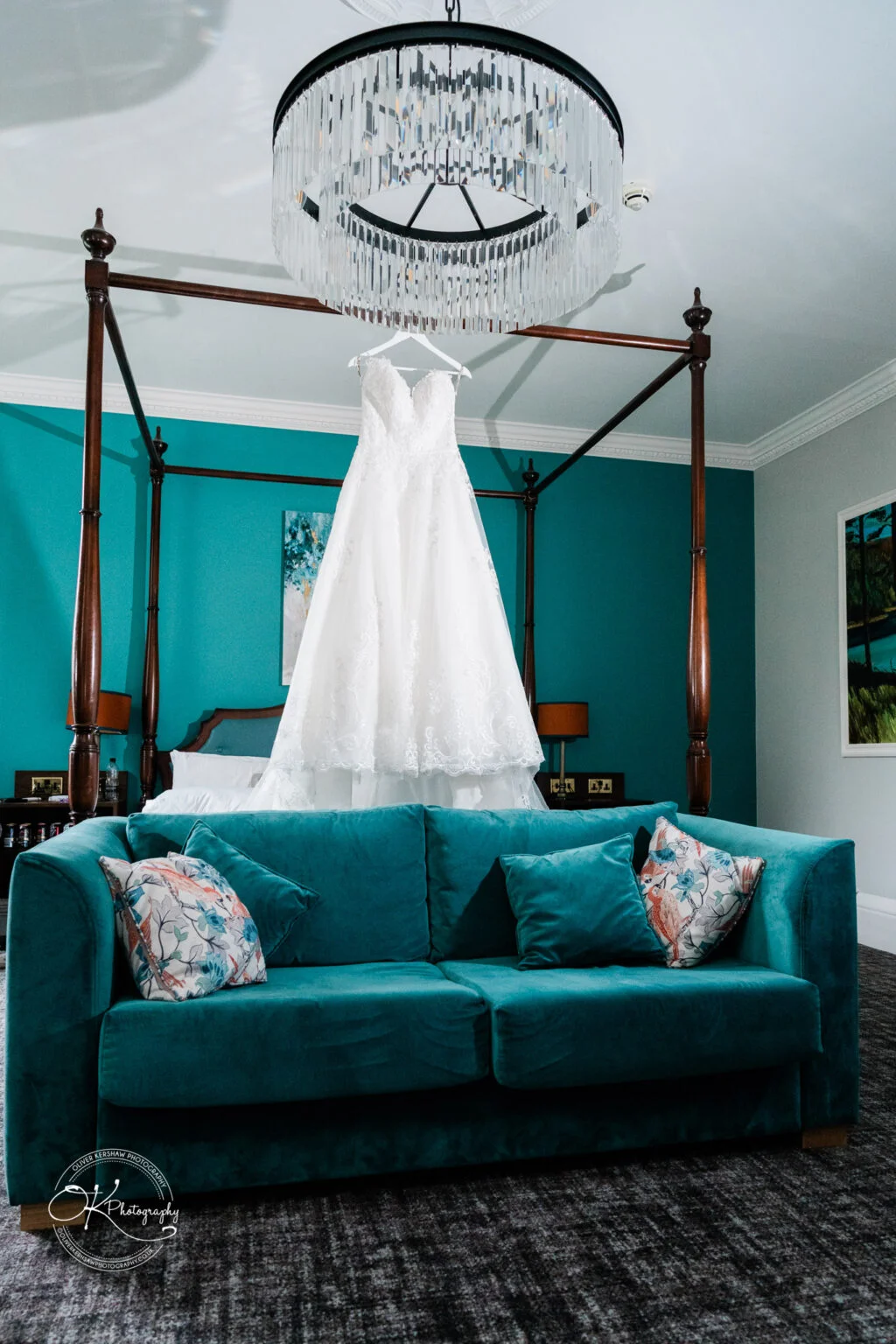 Makeney Hall Hotel - Wedding Photography A teal room with a teal sofa and a floral pillow, featuring a four-poster bed with a white wedding dress hanging, and a modern chandelier above.